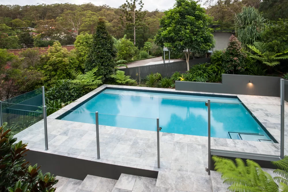 Modern swimming pool with a glass fence on the tile floor covered with a garden with green trees, there is a jungle from a distance with some house through it